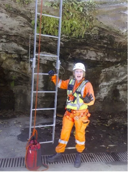 Veronica Hayes in high visibility overalls and helmet standing next to a ladder mounted to a rockface